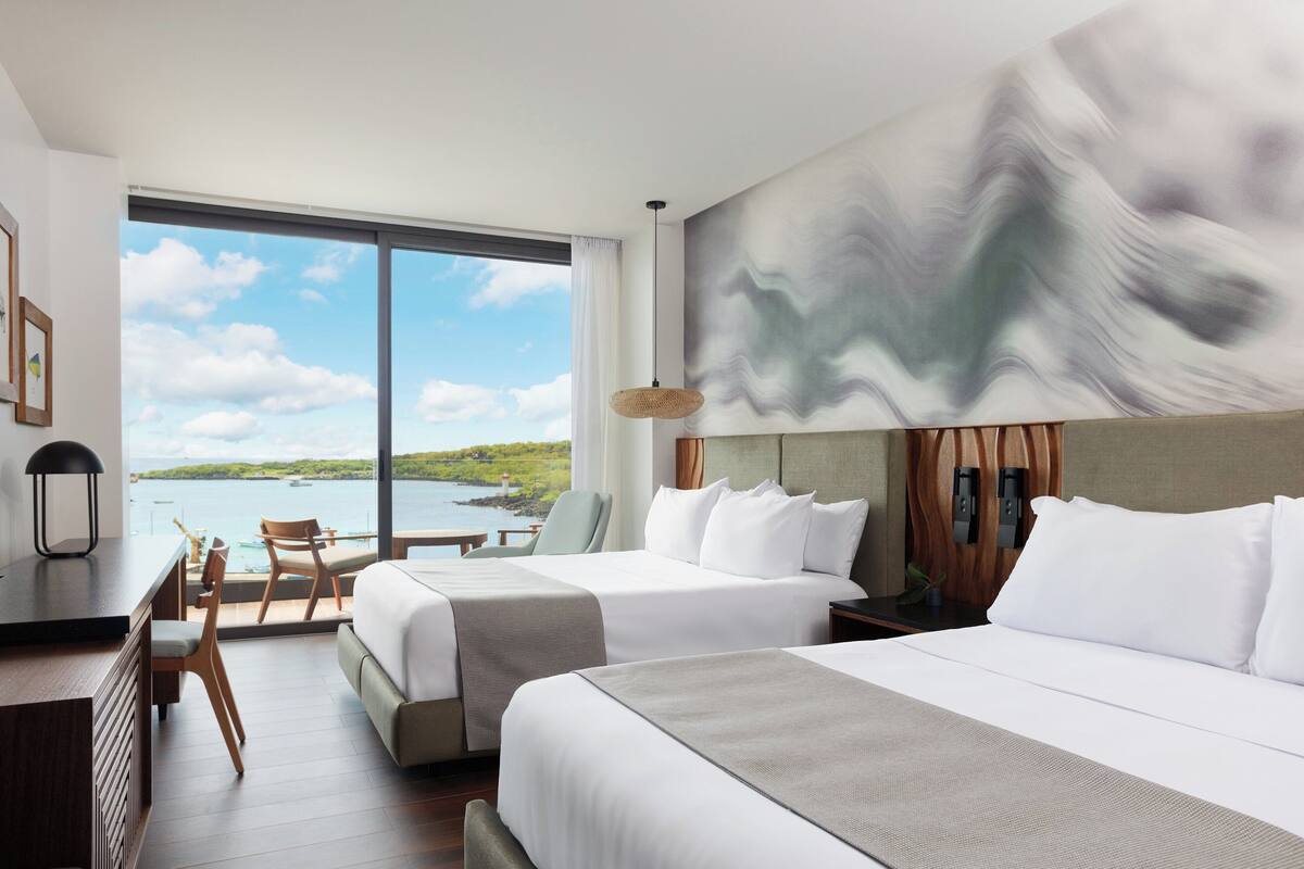 Une chambre d’hôtel avec deux lits, un bureau et une chaise. Une grande fenêtre offre une vue sur la mer et le paysage environnant.