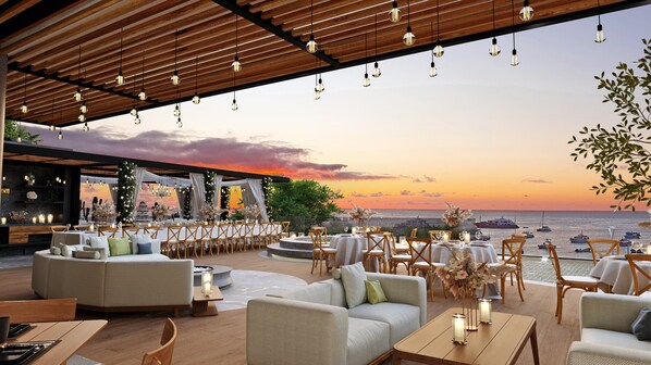 Un espace de restauration en plein air avec des tables et des chaises, un plafond en bois et une vue sur la mer au coucher du soleil.