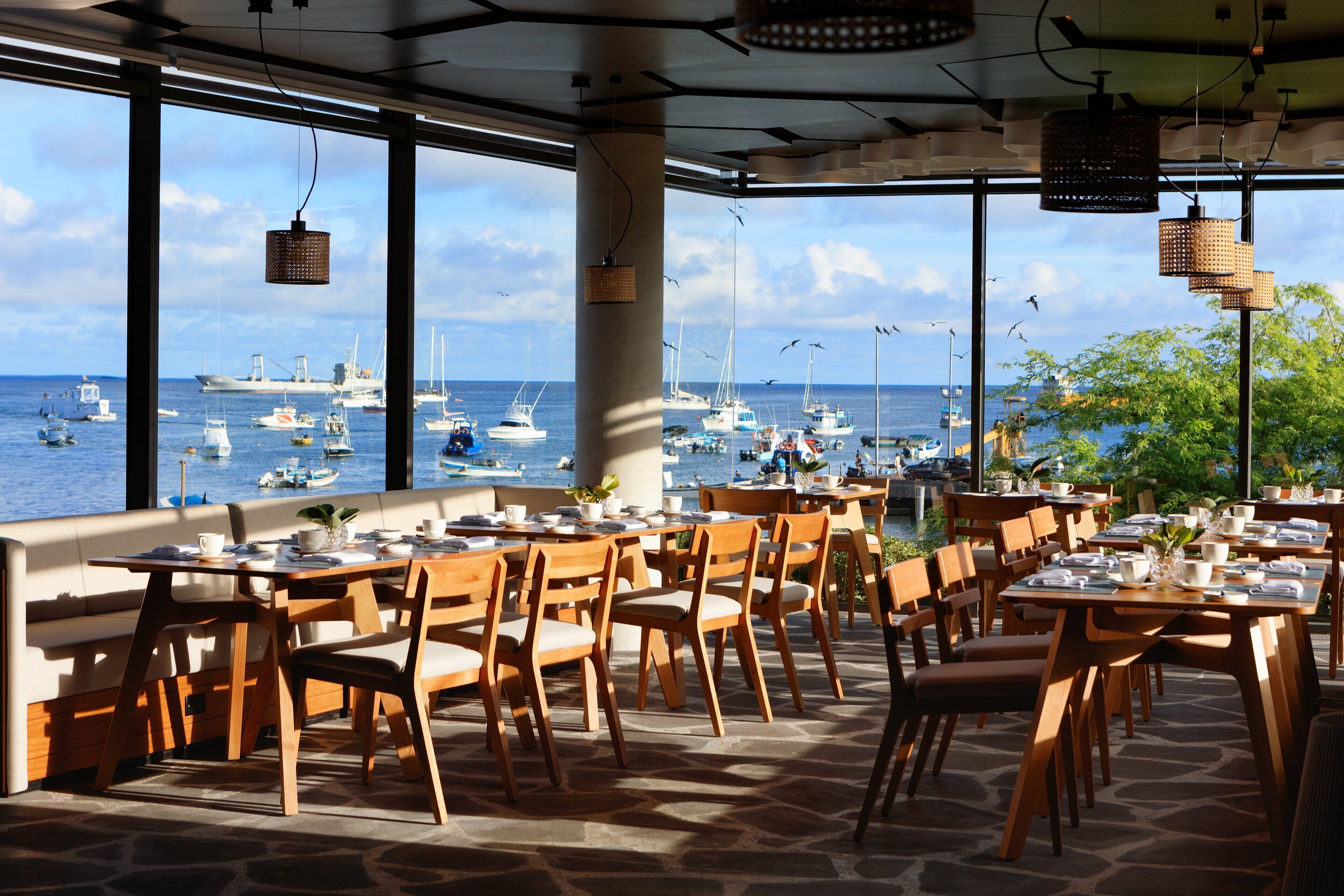 Un espace de restauration en plein air avec des tables et des chaises en bois, de grandes fenêtres donnant sur une marina où sont amarrés des bateaux.