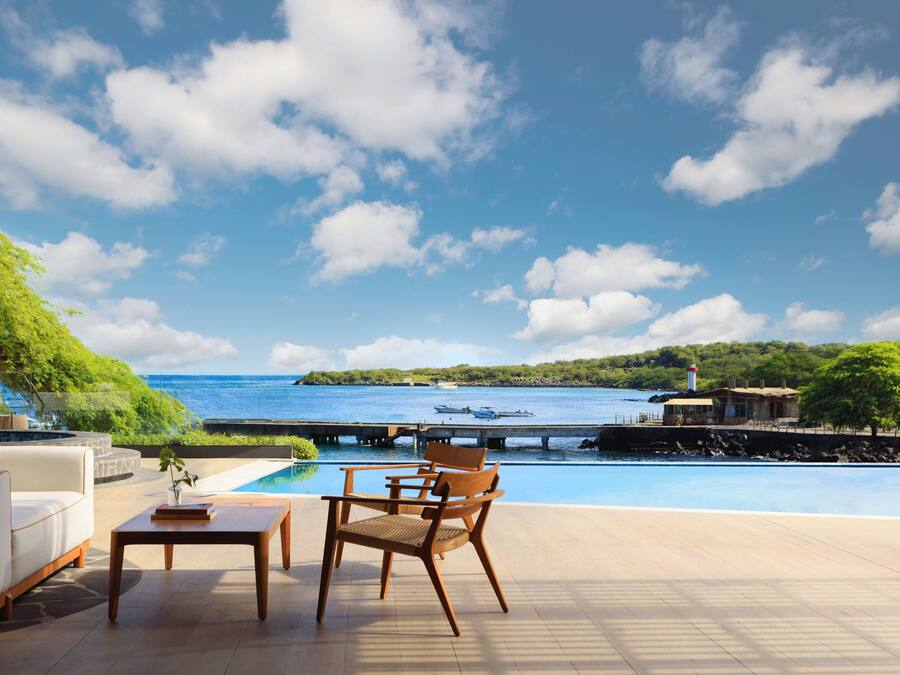 A poolside area with a view of the sea, a wooden chair, a sofa, and a small table.