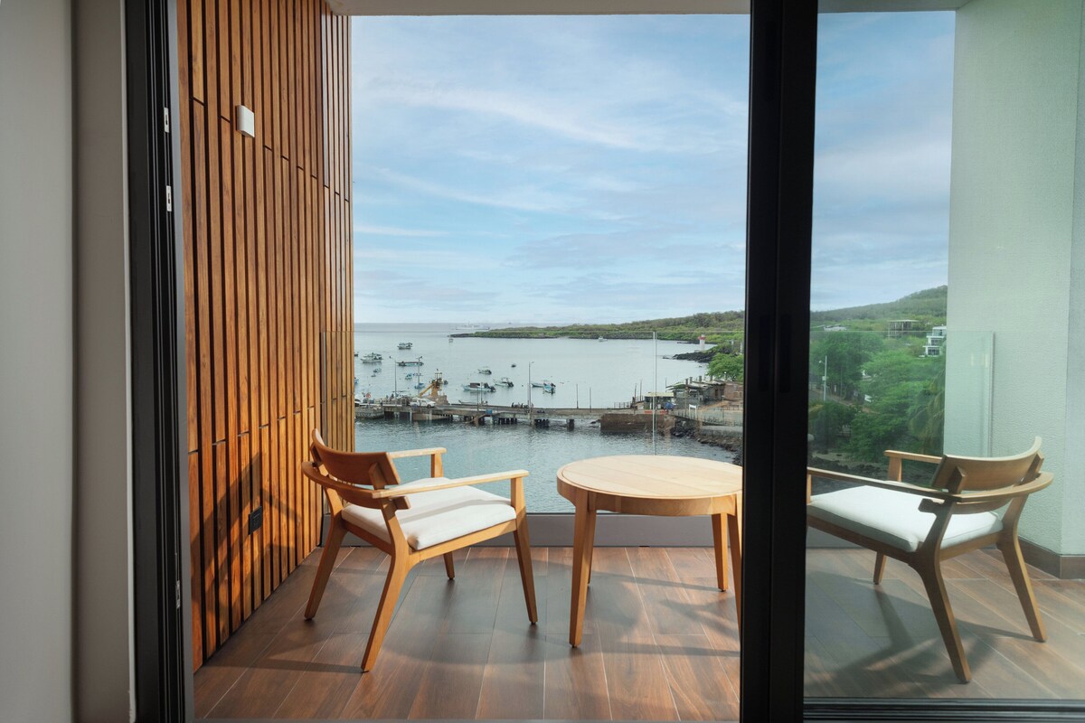 Ein Balkon mit Holzboden, zwei Holzstühle und ein runder Tisch. Durch die Glasüren ist eine Marina mit Booten und einer entfernten Küstenlinie zu sehen.
