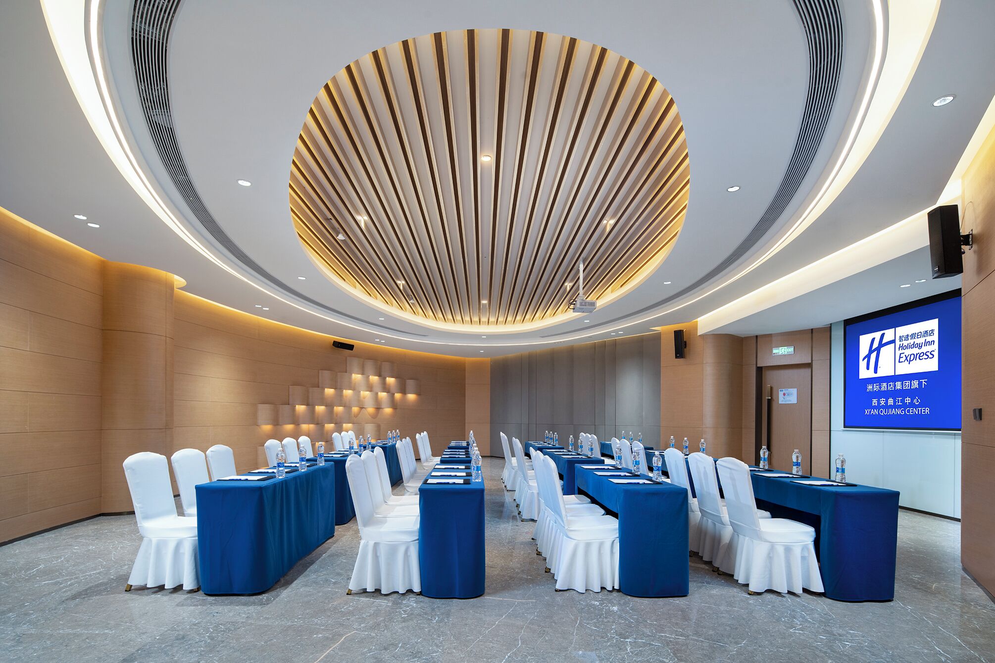 Un salón de conferencias con filas de mesas y sillas, un diseño moderno de techo y una pantalla grande que muestra información del hotel.