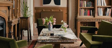 A room with a fireplace, a large bookcase, a marble coffee table, and green upholstered chairs.