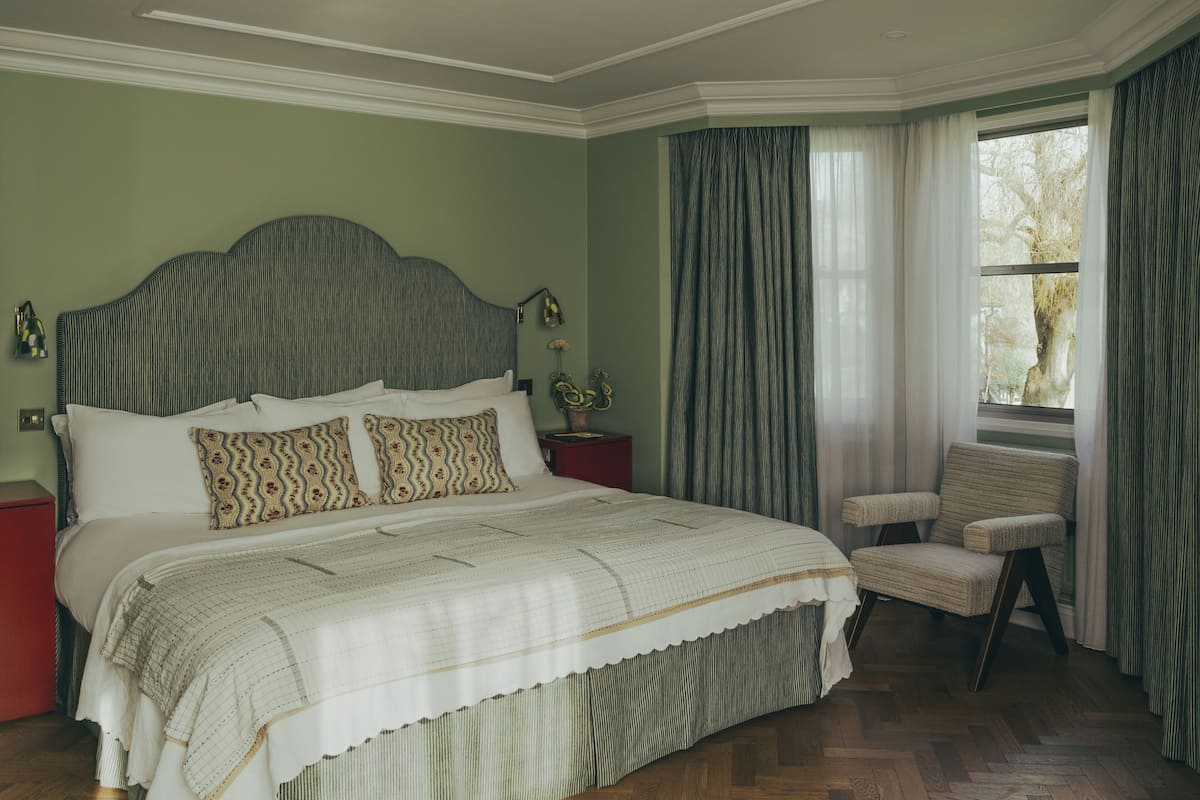 Quarto com cama grande, uma cadeira, mesa de cabeceira e janela com cortinas.