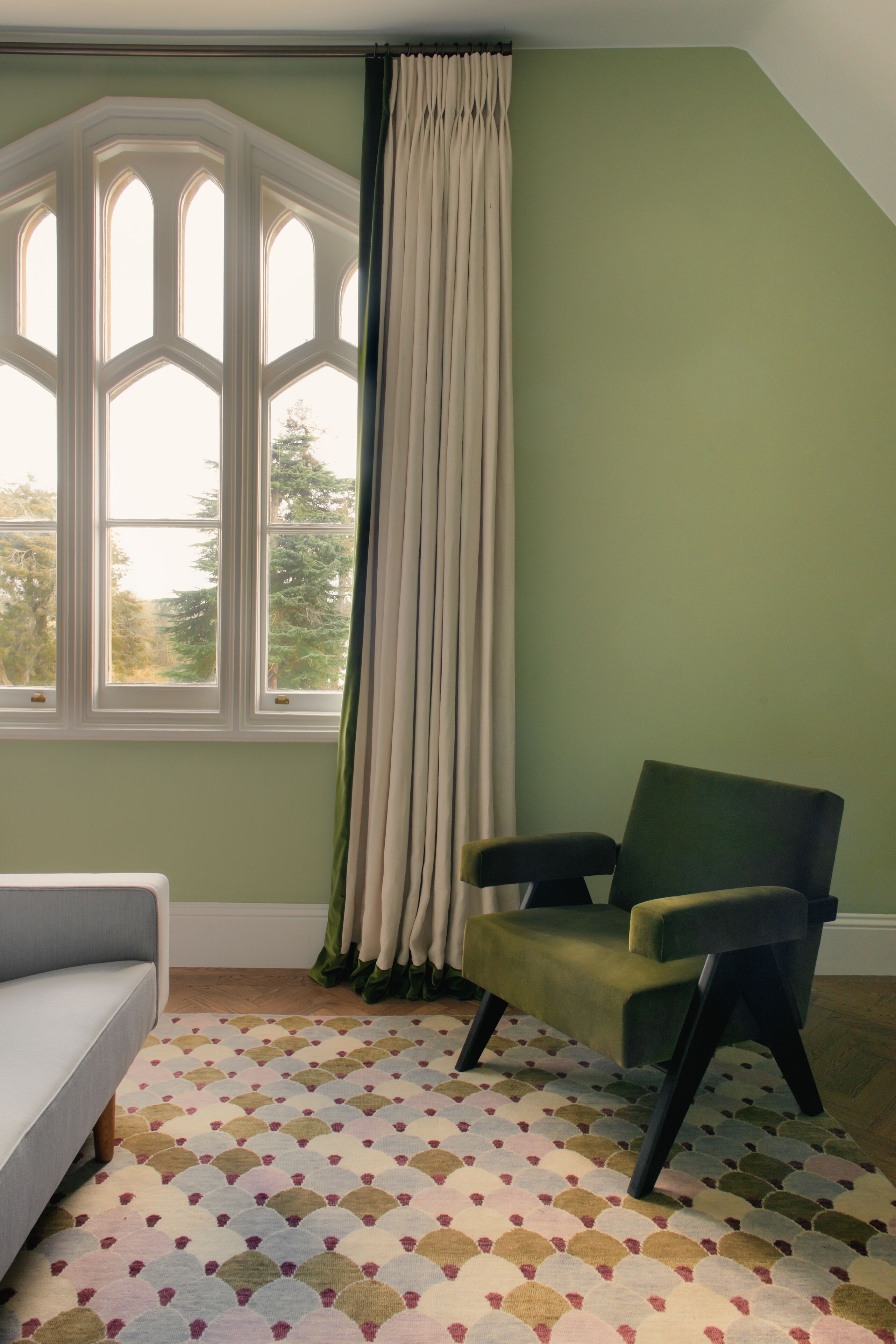 Una habitación con un ventanal grande, un sillón verde, un sofá blanco y una alfombra estampada.