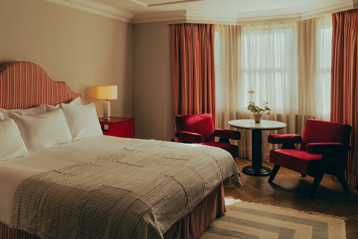 Quarto de hotel com uma cama grande, duas poltronas vermelhas, uma mesa redonda pequena com um vaso e uma luminária em um criado-mudo vermelho.