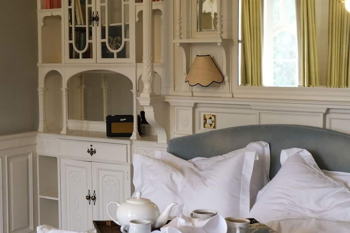 Quarto de hotel com uma cama grande, uma mesa de cabeceira com jogos de chá, um guarda-roupa com várias portas e um espelho sobre a cama.