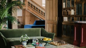 A cozy living room with green sofas, a coffee table with books and a chess set, a large plant, and a wooden staircase leading to another room. - Oakley Court (Windsor)