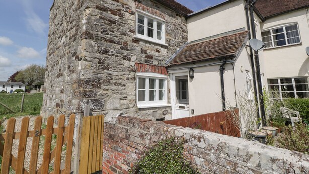A two-story stone building with a white door and windows, a brick wall, and a wooden gate. - 2 Court Farm (Wareham)