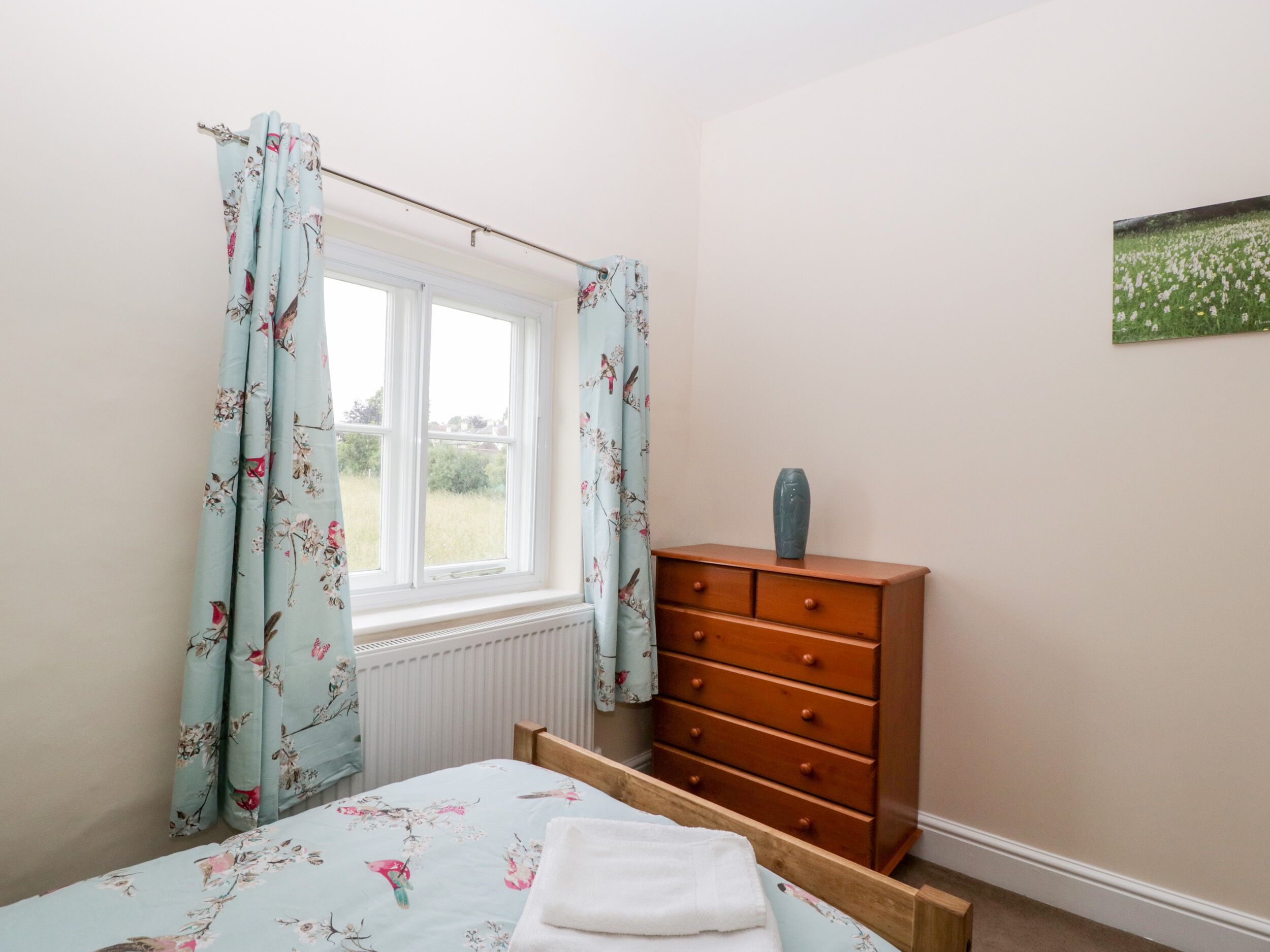 A bedroom with a bed, a dresser, a window with floral curtains, and a radiator.