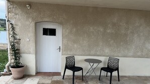 A covered patio with a white door, two black chairs, a small round table, and a potted plant. - Gîte des oliviers (Vénéjan)