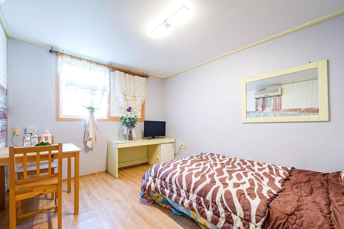 A bedroom with a bed, a desk, a chair, a TV, and a window with curtains.