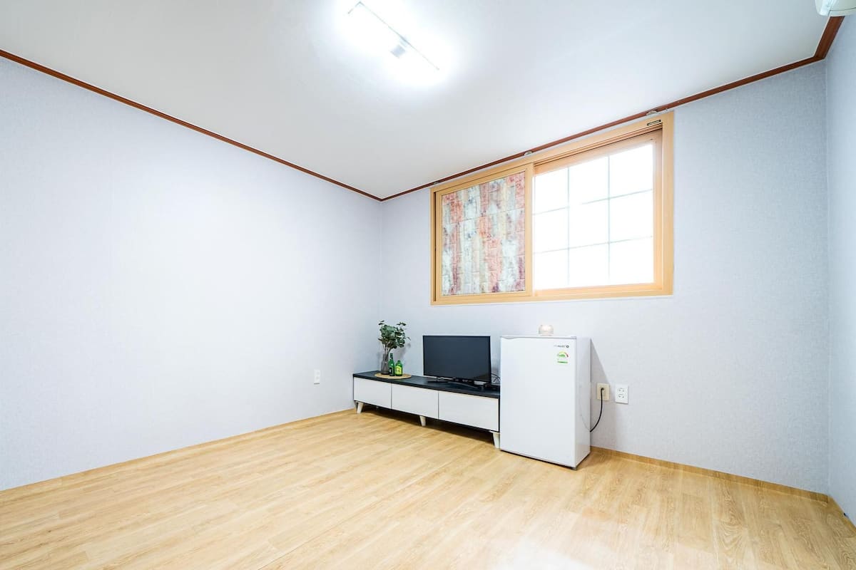 A room with a wooden floor, a desk with a computer, a small refrigerator, and a window with curtains.