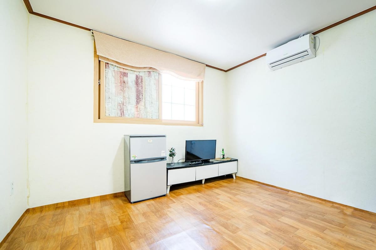 A room with a wooden floor, a desk with a computer, a small refrigerator, a window with curtains, and an air conditioning unit.