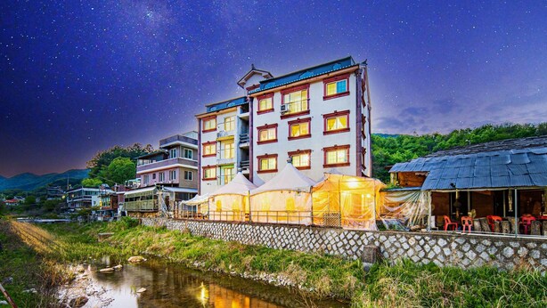 A multi-story building with a stone wall and a river in the foreground. - Gangchon Pine Hill Pension (Chuncheon)