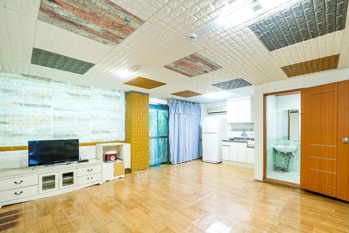 A spacious room with a wooden floor, a television, a small cabinet, a kitchen area with a refrigerator, and a bathroom with a sink and mirror.