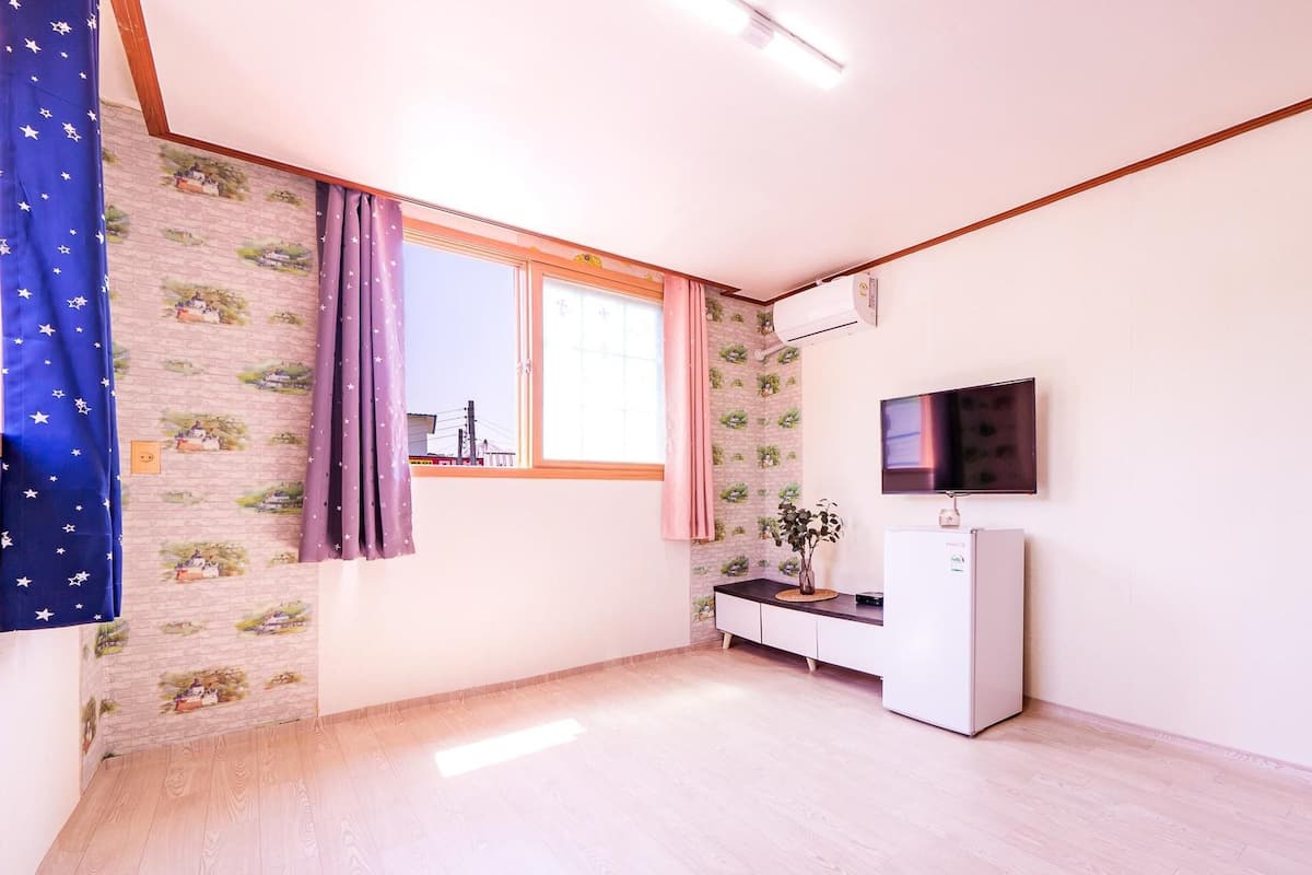 A small, well-lit room with a window, a TV mounted on the wall, a white cabinet, and a wooden door.