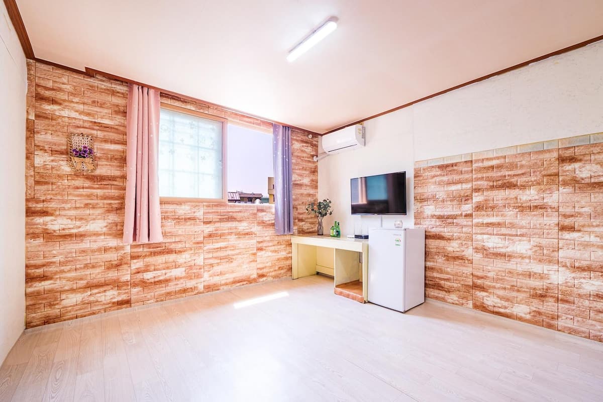 A small room with a brick-patterned wall, a desk with a computer, a TV, and a refrigerator.