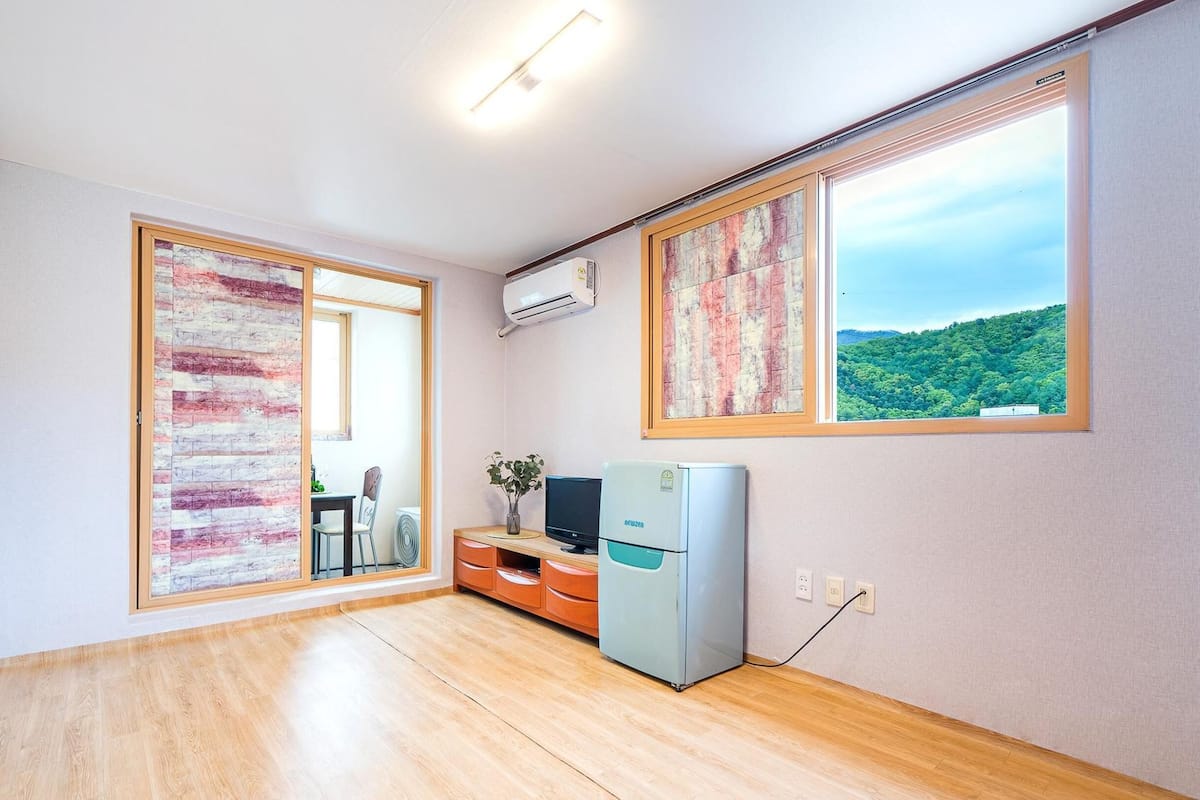 A room with a wooden floor, a desk with a computer, a small refrigerator, and a window with a view of a forest.