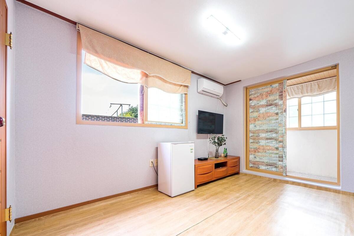 A room with a wooden floor, a small white refrigerator, a TV mounted on the wall, and a sliding door with a view of the outside.