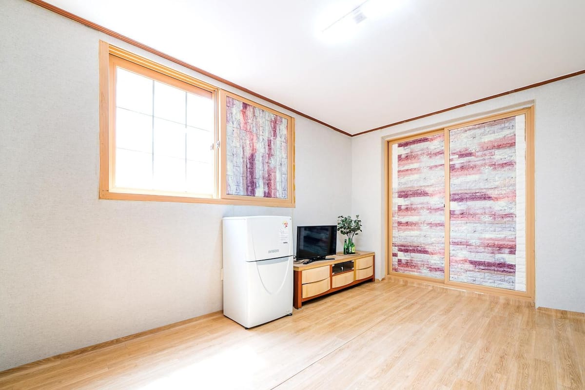 A room with a wooden floor, a white refrigerator, a wooden cabinet with a television, and a sliding door with a pink and white abstract design.