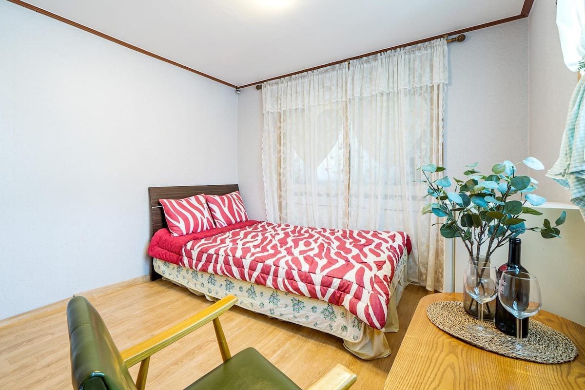 A bedroom with a bed, a chair, a table with a vase and glasses, and a window with curtains.