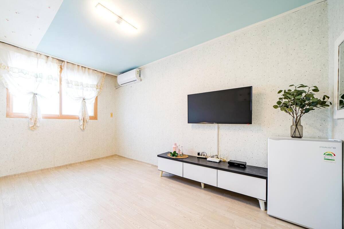 A modern, minimalist living room with a flat-screen TV mounted on the wall, a white refrigerator, a wooden cabinet, and a large window with sheer curtains.