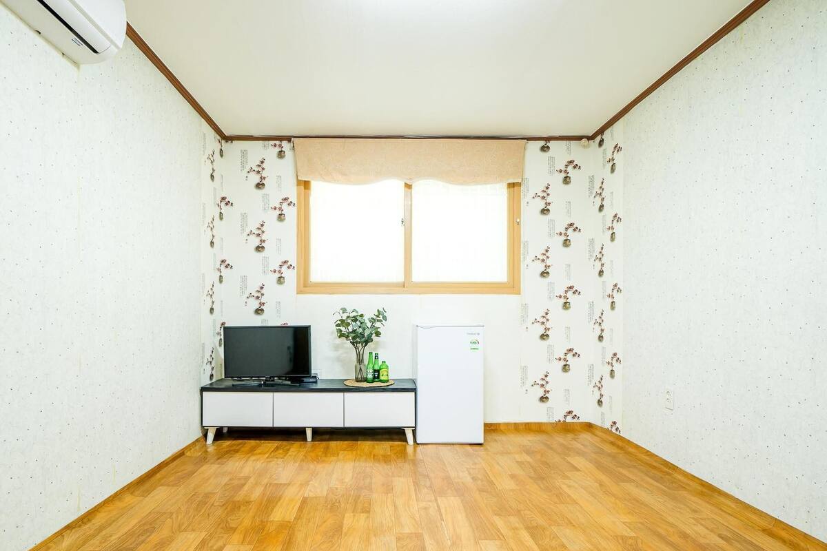 A room with a wooden floor, a desk with a computer, a small refrigerator, and a window with floral wallpaper.