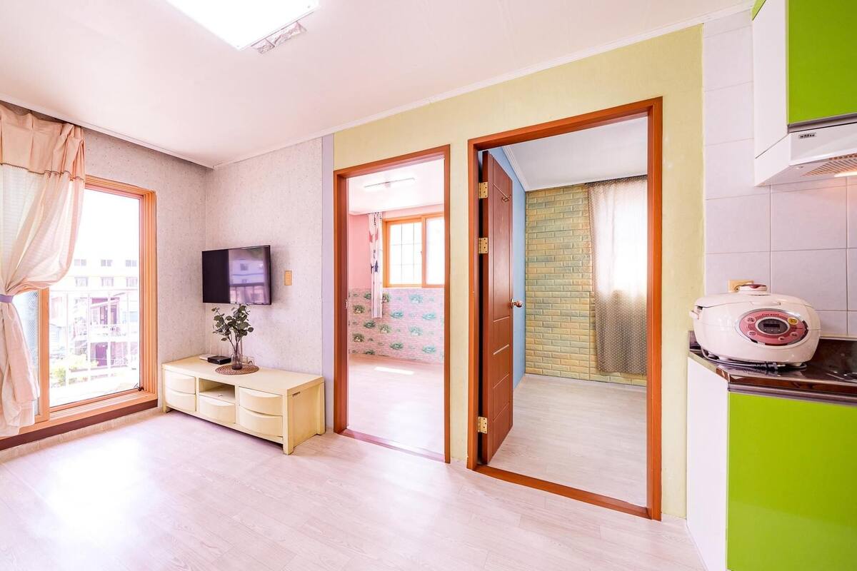 A modern apartment with a large window, a flat-screen TV, a small desk, and a kitchen area with a rice cooker.