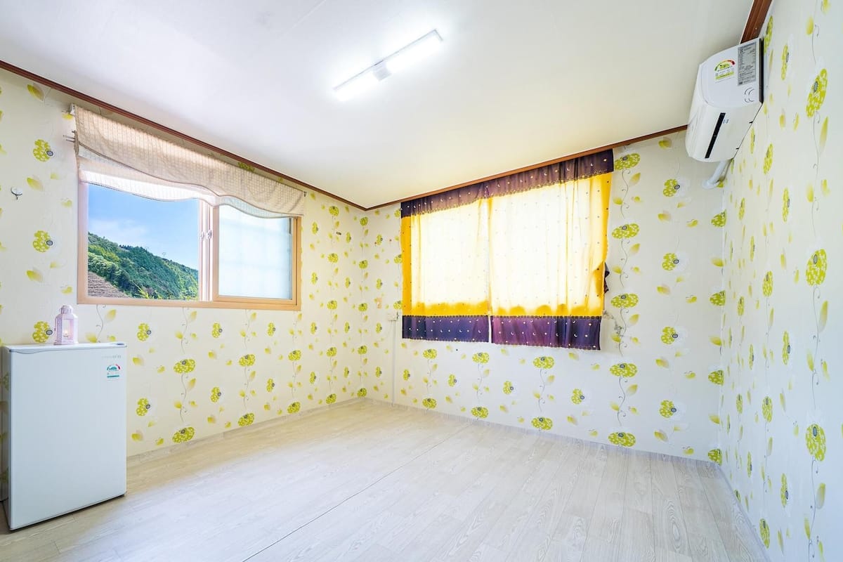 A room with floral wallpaper, a window with curtains, a white refrigerator, and an air conditioning unit.