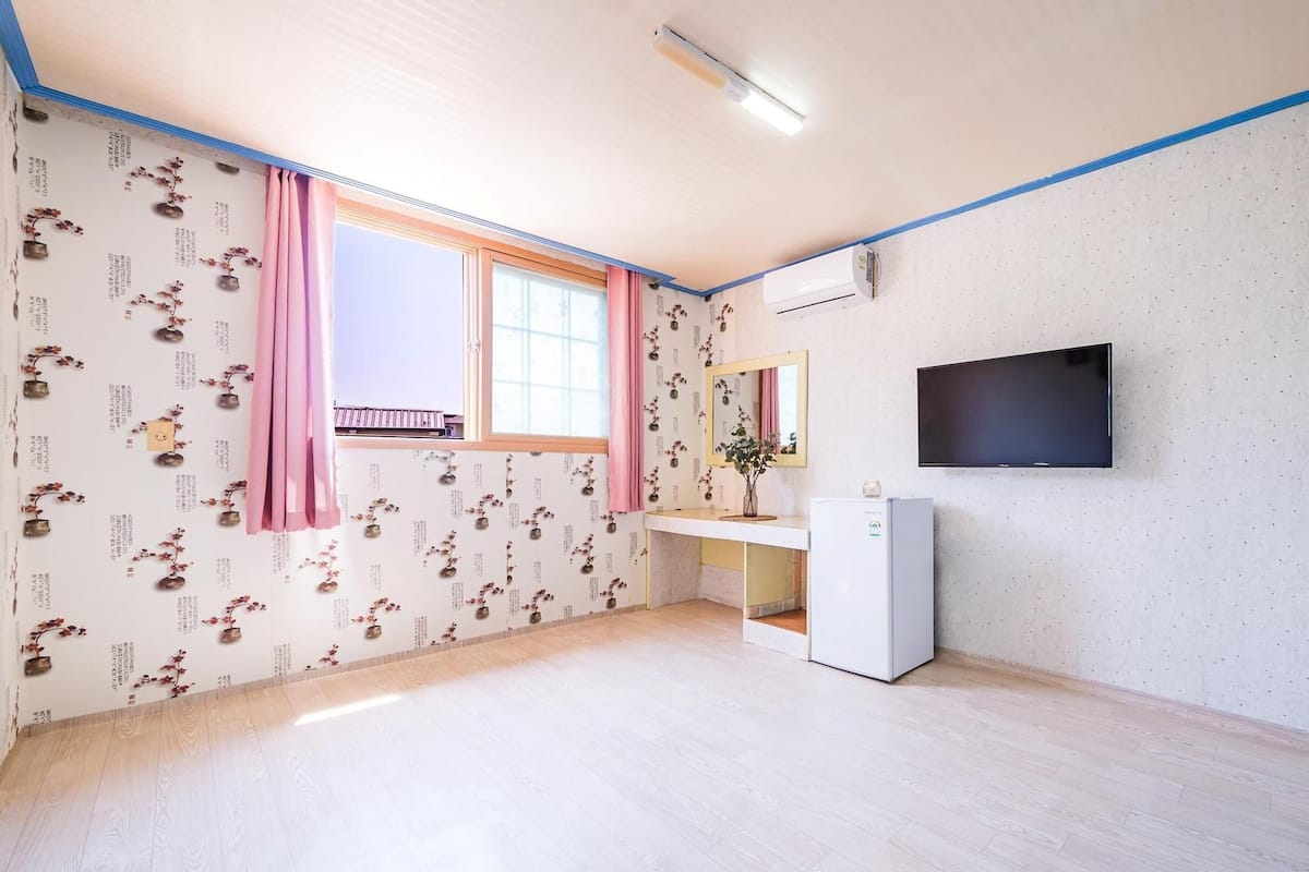 A room with a large window, a flat-screen TV mounted on the wall, and a white desk with a mirror.
