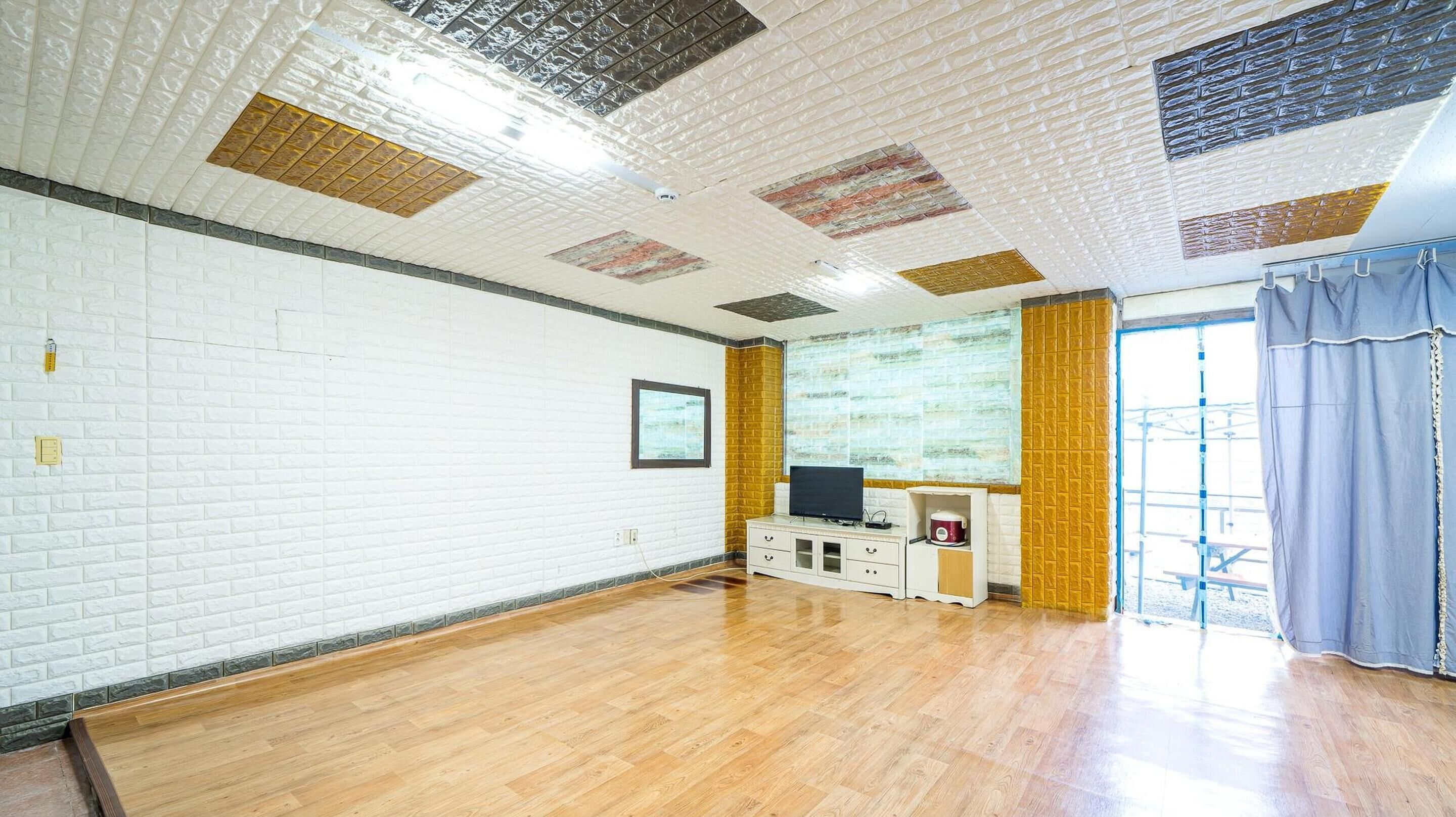 A spacious room with a white brick wall, wooden flooring, a flat-screen TV, a small cabinet, and a large window with curtains.