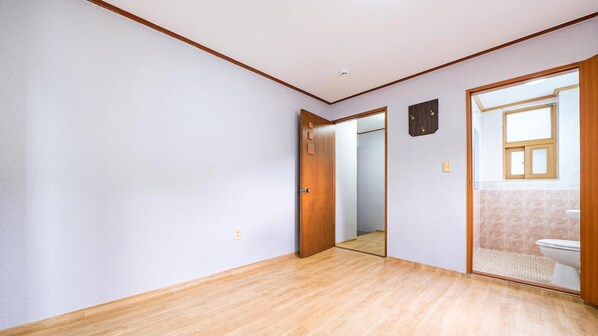 Una habitación con suelo de madera, paredes blancas, una puerta de madera y vista al baño, que tiene inodoro y ventana.