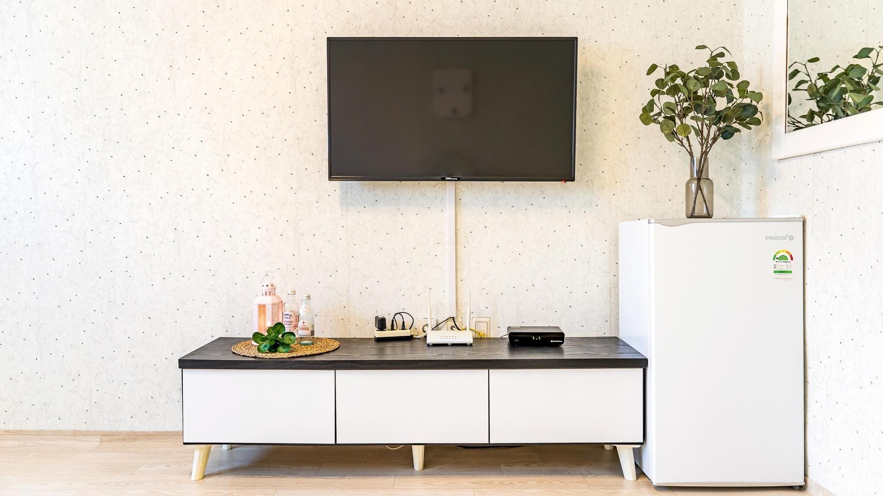Un salon moderne avec un téléviseur à écran plat fixé au mur, un réfrigérateur blanc, un meuble en bois surmonté d’un tapis tressé et une plante en pot.