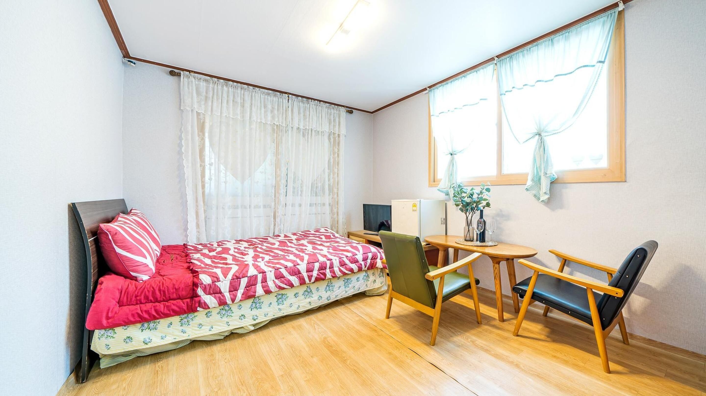 A small, cozy room with a bed, a desk, a chair, and a window with sheer curtains.