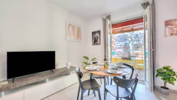 A modern living room with a flat-screen TV, a dining table with chairs, a potted plant, and a balcony with a view.