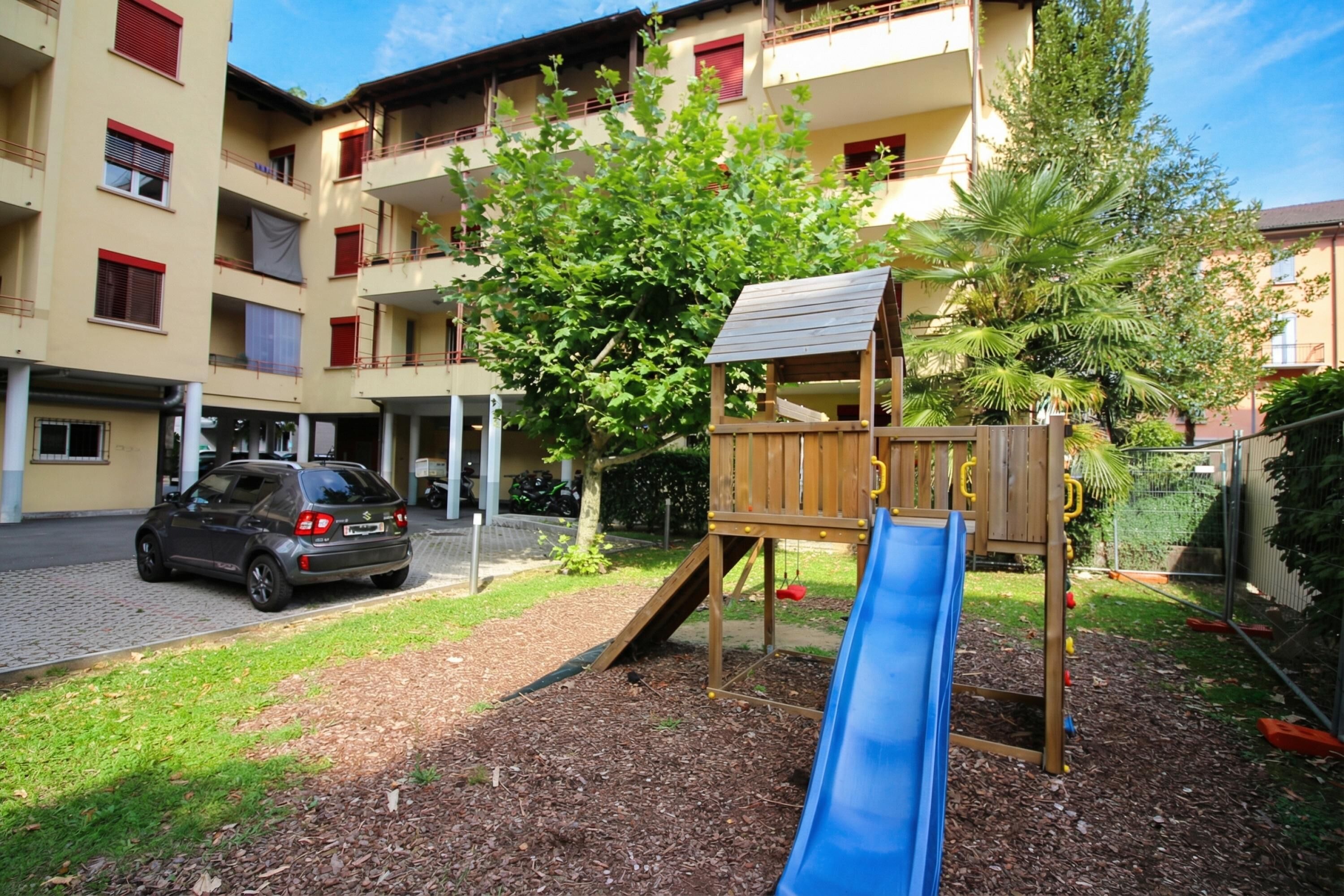 A playground with a slide and climbing structure, surrounded by a building with balconies and a parked car.