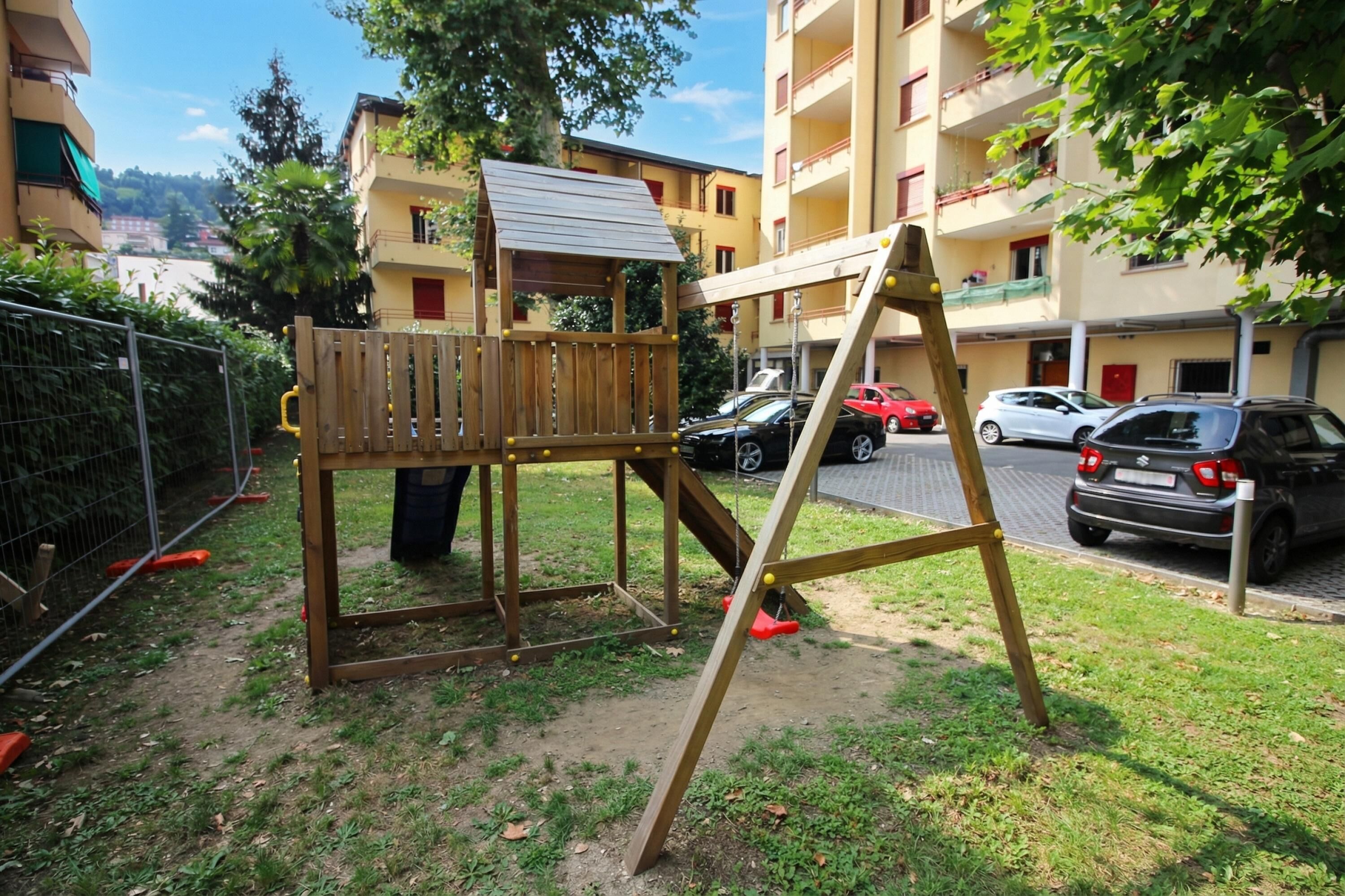 Un parque infantil con una estructura de madera y columpios en una zona residencial, con coches aparcados y edificios al fondo.