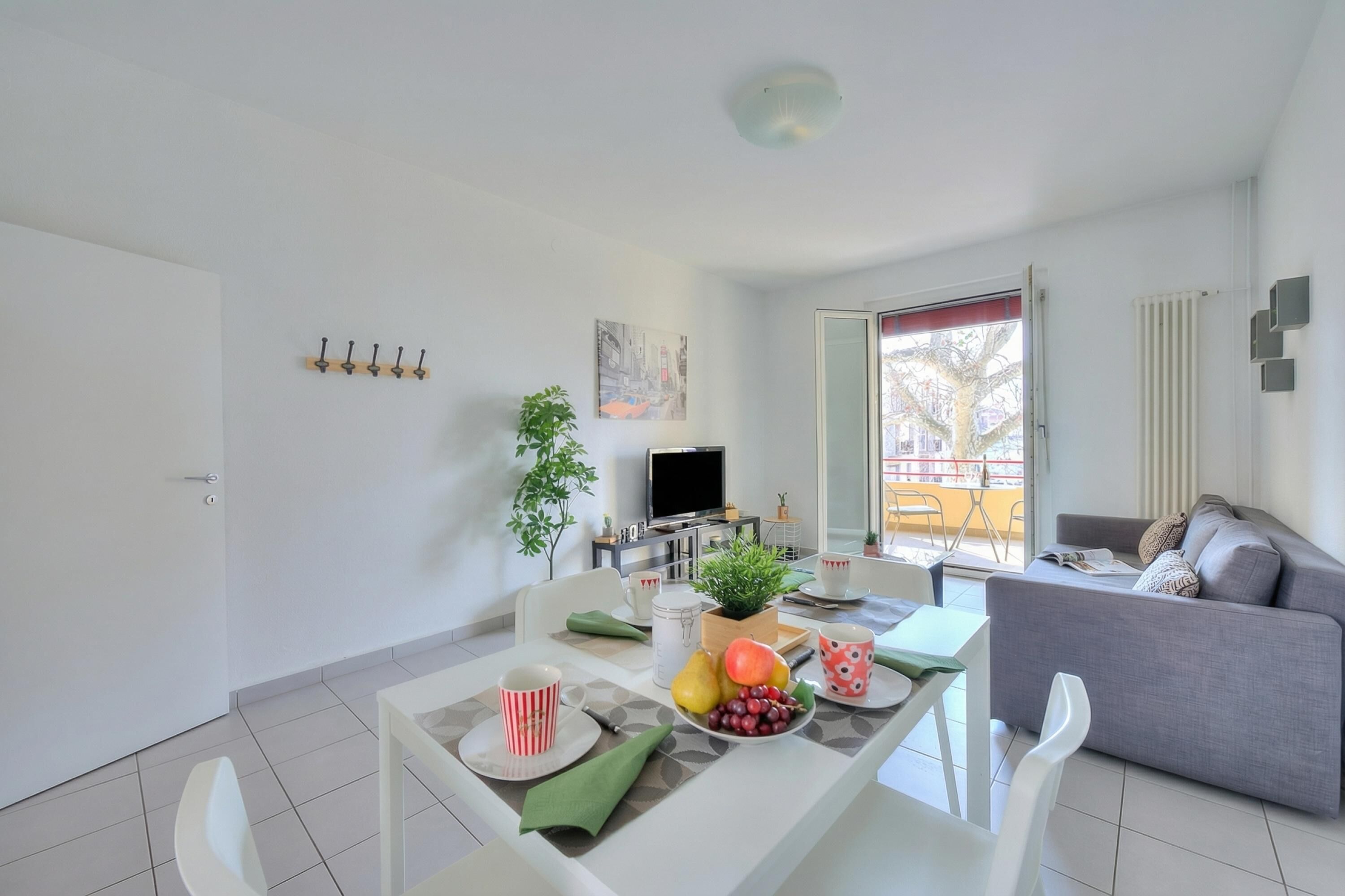 A modern living room with a grey sofa, a dining table with fruit and cups, a TV, and a balcony with a view.