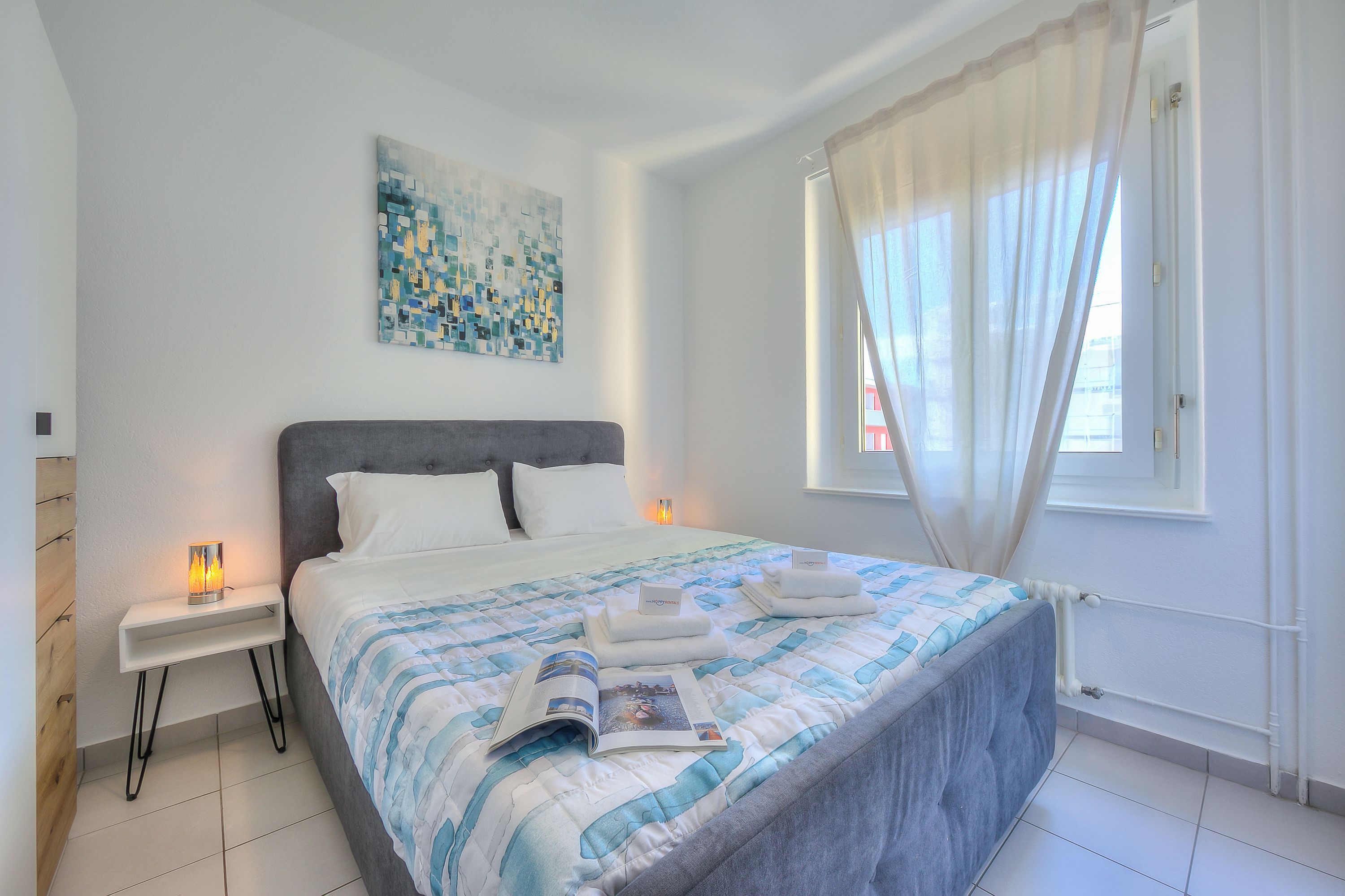 A modern bedroom with a large bed, white nightstands, a bedside lamp, a wall art piece, and a window with sheer curtains.