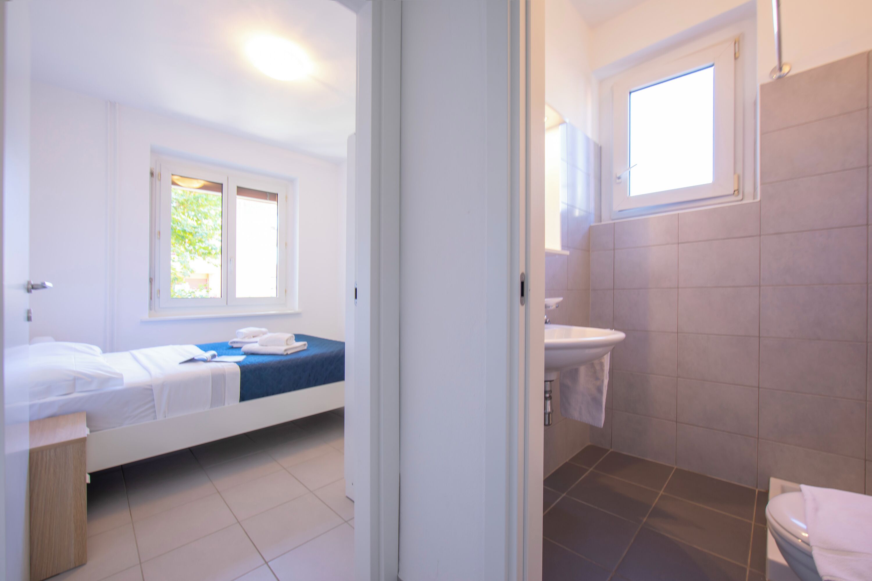 A bedroom with a single bed, a window, and a bathroom with a sink and toilet.