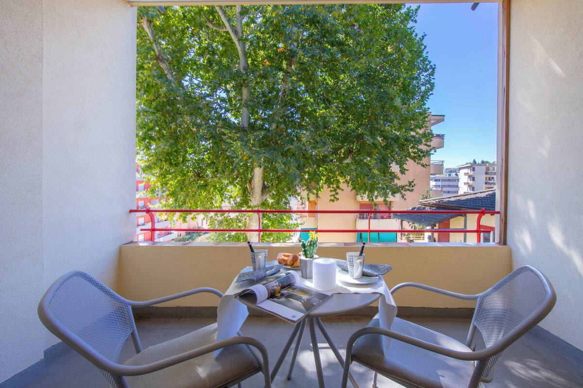 Un balcón con una mesa preparada para dos, una silla y vistas a árboles y edificios.