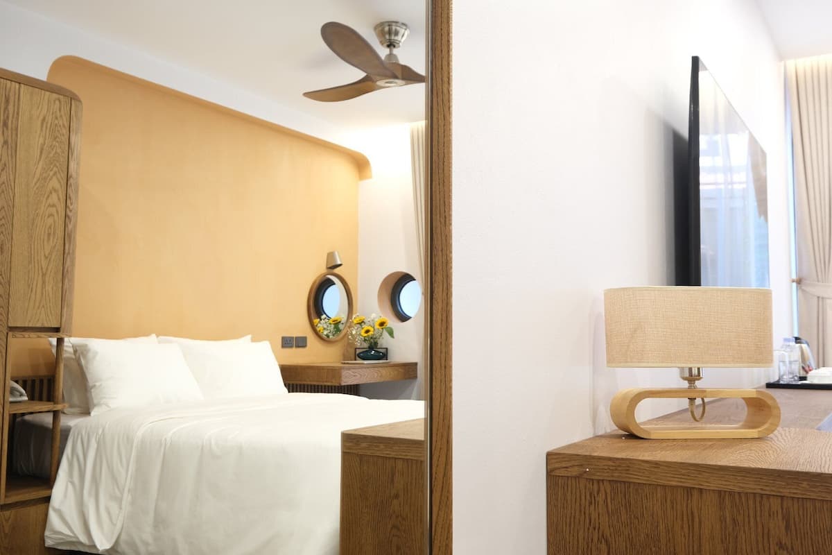 A hotel room with a bed, a wooden headboard, a ceiling fan, a mirror, a lamp, and a window with curtains.