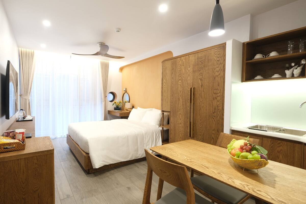 A hotel room with a bed, a desk, a chair, a small table with fruit, a mirror, and a window with curtains.