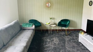 A living room with a grey sofa, two green velvet armchairs, a small round table with a vase, and a TV stand. - Lady Wilson Apartment (Airdrie)