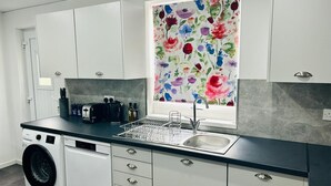 A modern kitchen with white cabinets, a black countertop, a built-in washing machine, and a window with a floral view. - Lady Wilson Apartment (Airdrie)