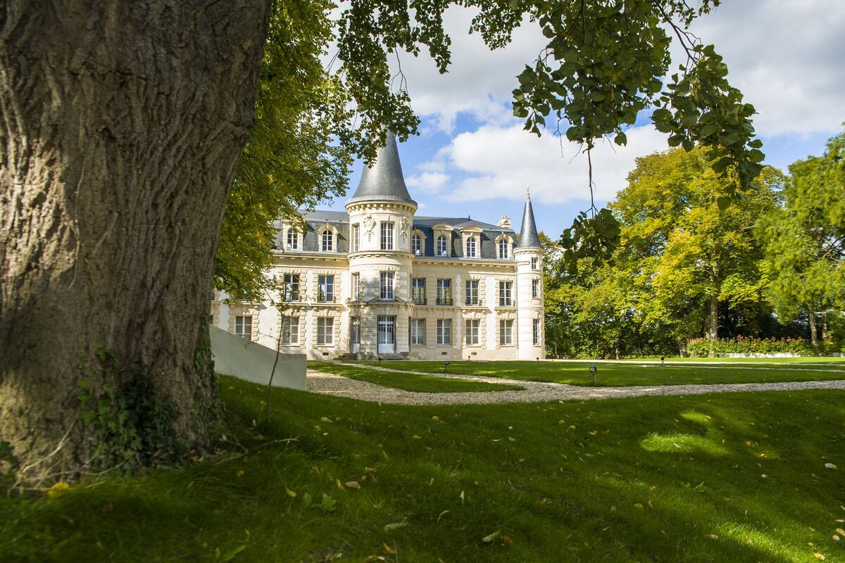 A large, historic building with a central tower and multiple wings, surrounded by a well-maintained lawn and trees.