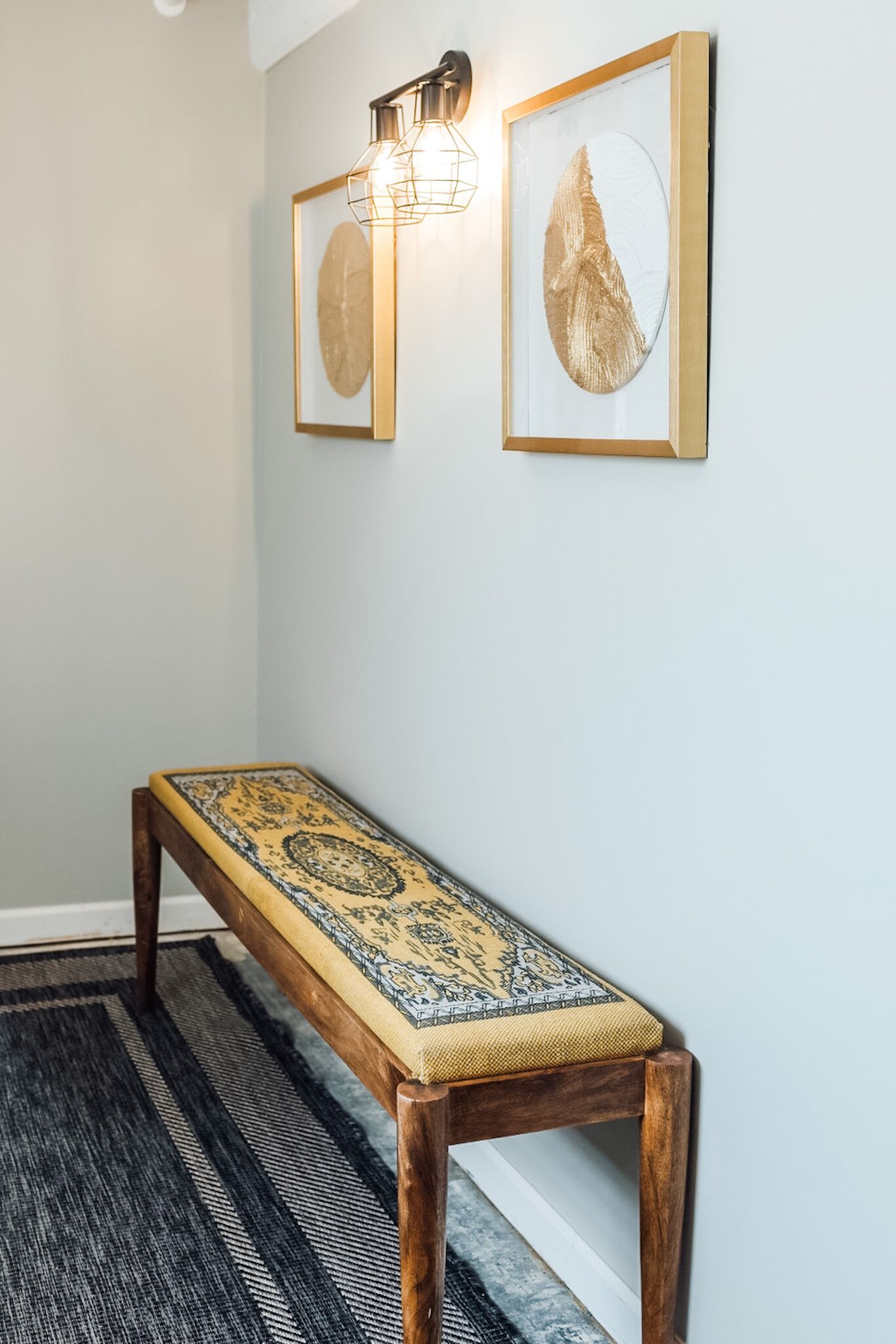 A bench with a patterned cushion, a wall-mounted light fixture, and two framed artworks.