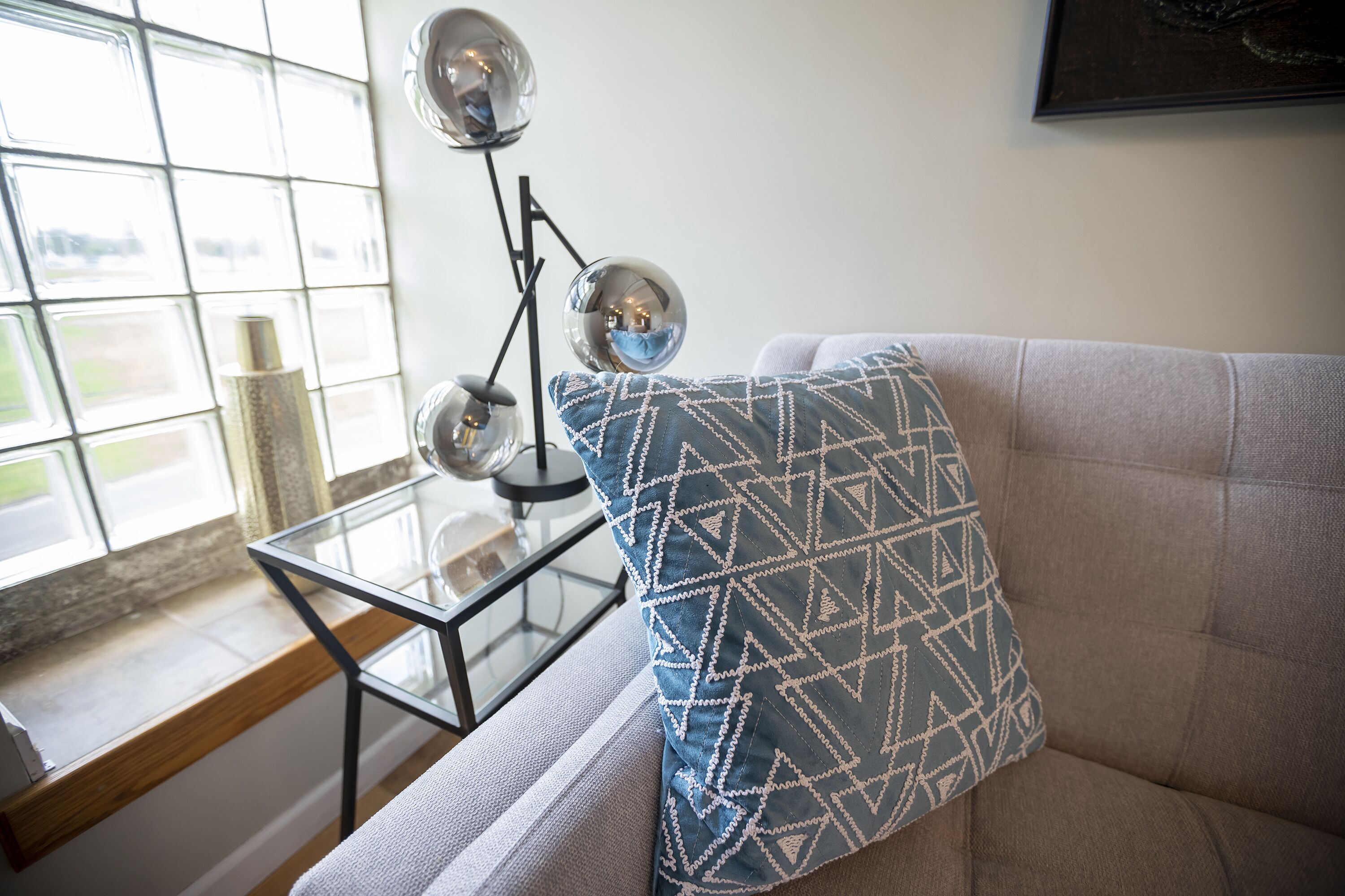 A living room with a sofa, a patterned pillow, a glass table, and a window with a view.