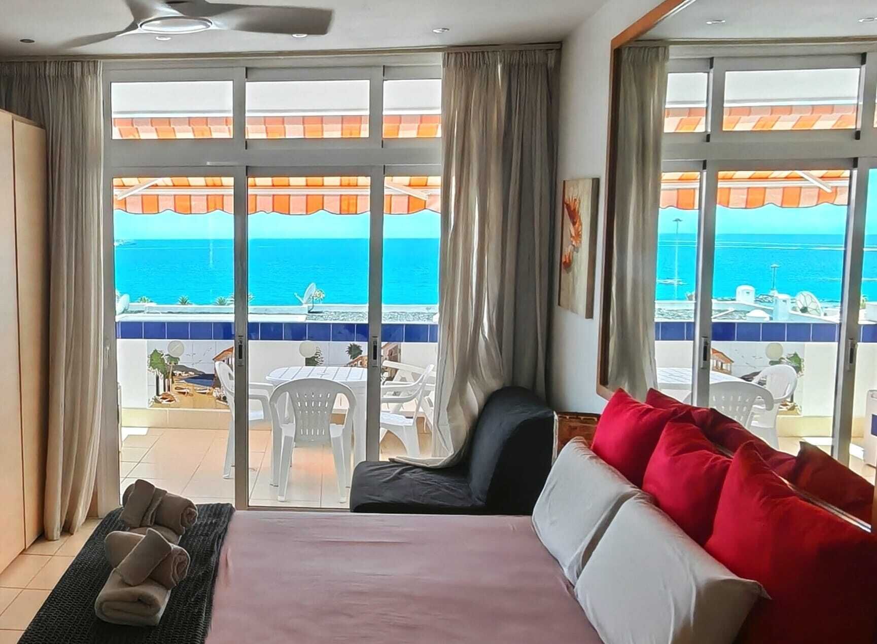 A hotel room with a large sliding glass door leading to a balcony with a view of the sea. There is a bed with red and white pillows, a chair, and a small table with a fan.
